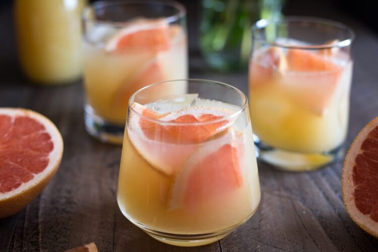 Three glasses of grapefruit juice with ice, displayed on a wooden table alongside grapefruit halves.